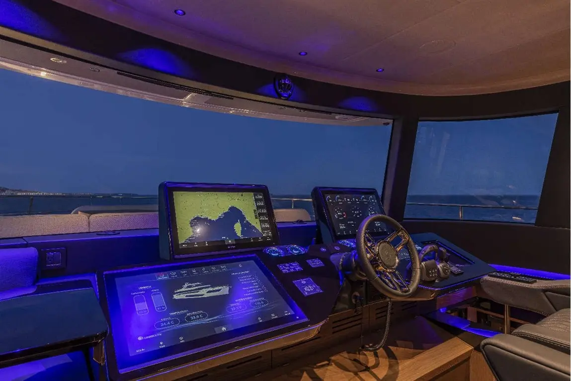 Modern yacht cockpit with navigation screens, steering wheel, and ambient lighting; view of water at dusk outside.
